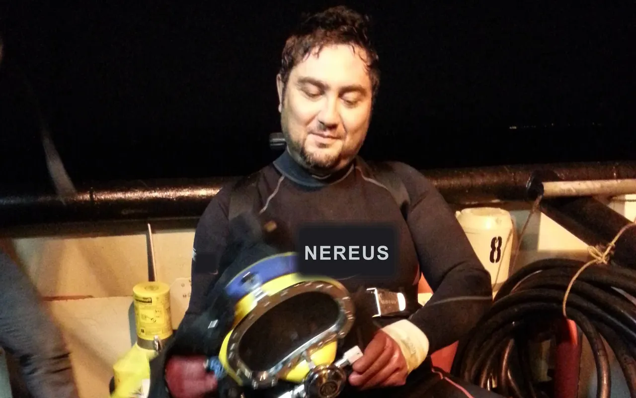 Commercial diver suited up on a workboat deck at night with a Kirby Morgan helmet in his lap